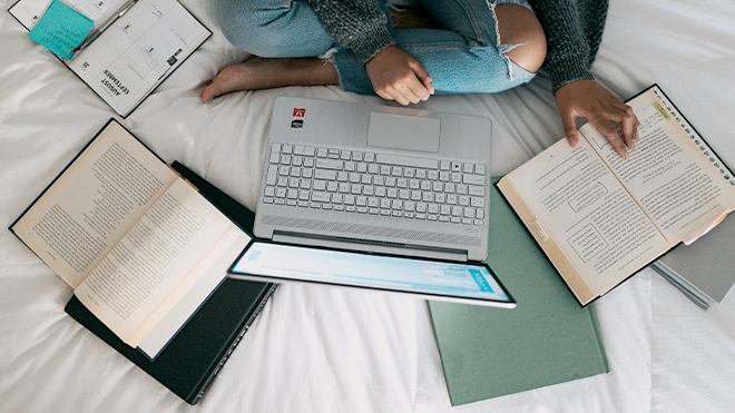 Person auf einem Bett sitzend mit Laptop und Büchern drum herum